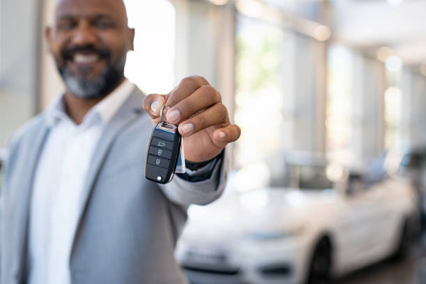 Smiling salesman holding new car keys in car showroom. Businessman is suit in car showroom showing car keys. Closeup hands of african man holding keys on car rental salon with copy space.