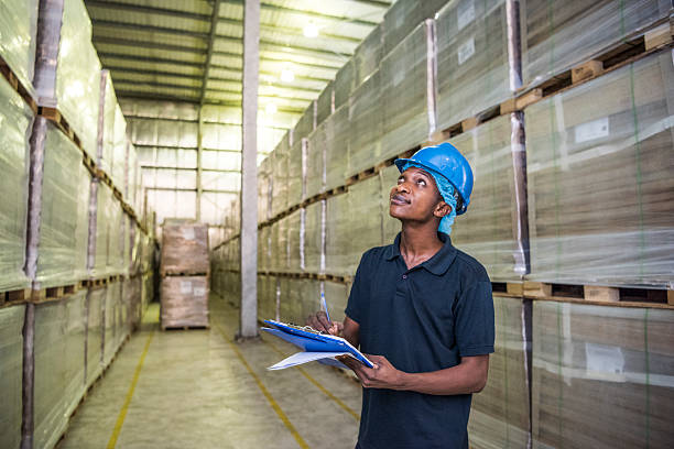 Ethnic industrial worker wearing hard hat holding clipboard, looking up at shelves in warehouse. Young male worker doing stock take, in factory warehouse.